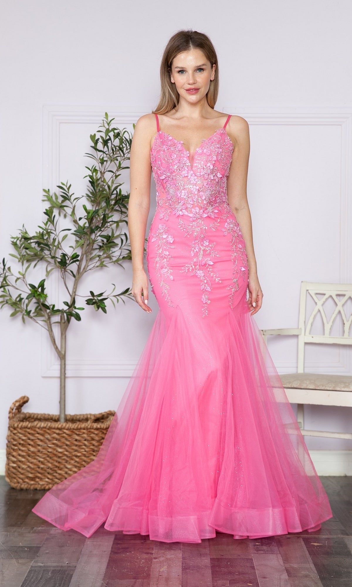 A woman stands indoors wearing the Sheer-Corset-Bodice Long Mermaid Prom Dress 9374, a fitted pink gown with floral embroidery and thin straps. A potted plant and white bench can be seen in the background.